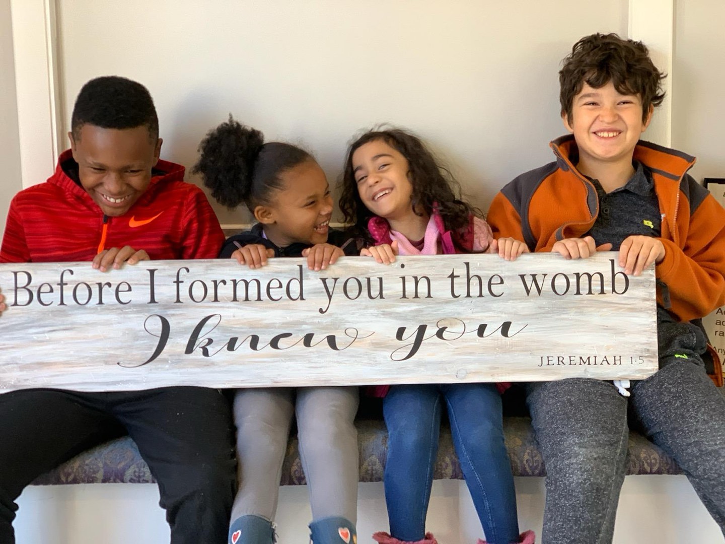 Four children sitting together at the center, holding a painted wooden sign that reads: Before I formed you in the womb I knew you. Jeremiah 1:5