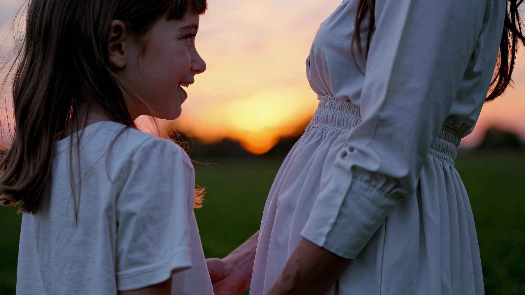 A young girl smiling up at her pregnant mother at sunset
